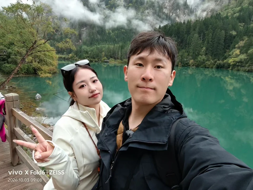 两人在九寨沟湖边合影，背景是青绿色湖水与云雾中的山林